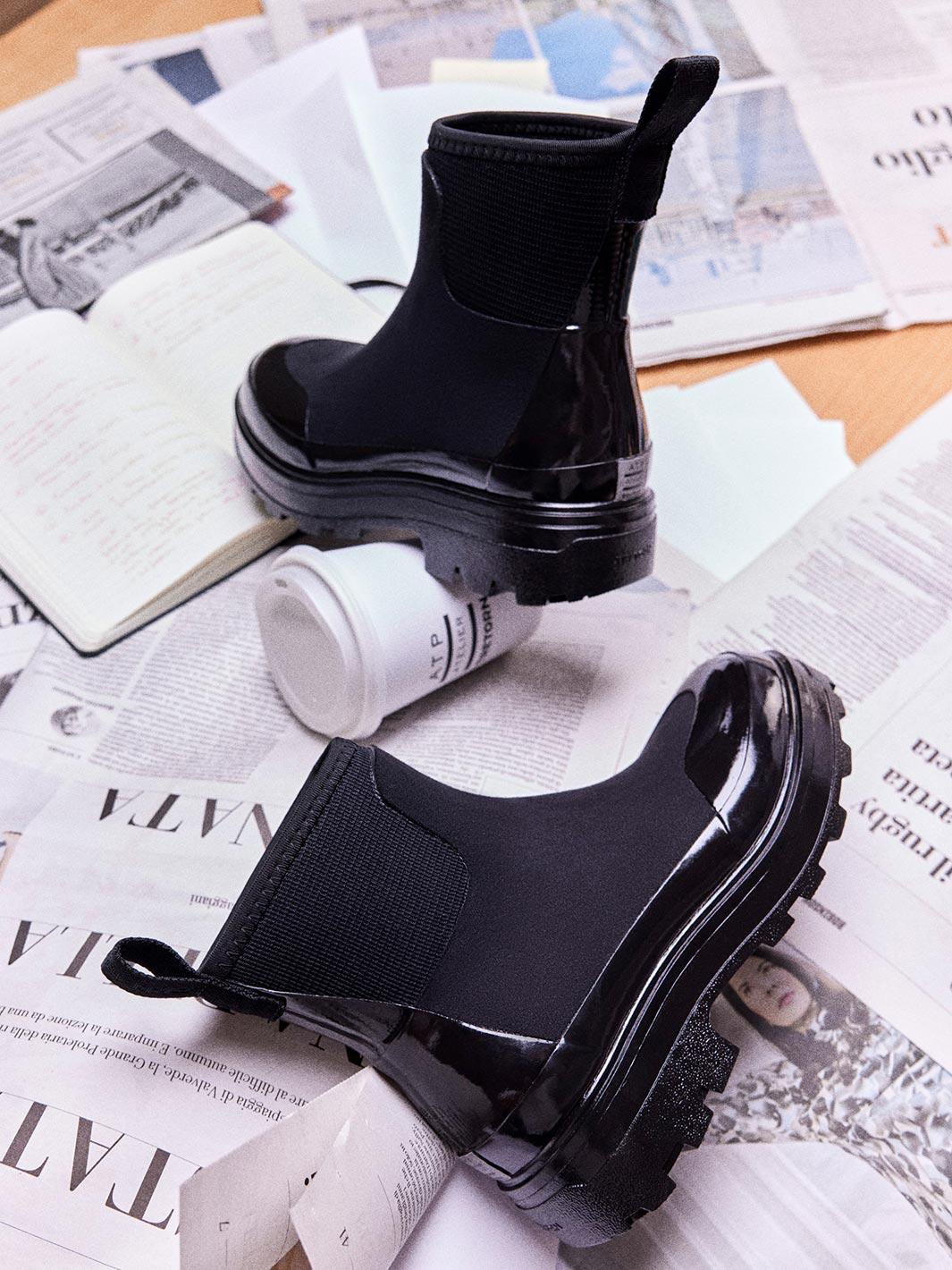 ATP Atelier Chelsea-style rubber boots with elastic side panels and chunky glossy soles styled on scattered newspapers next to a branded paper coffee cup.