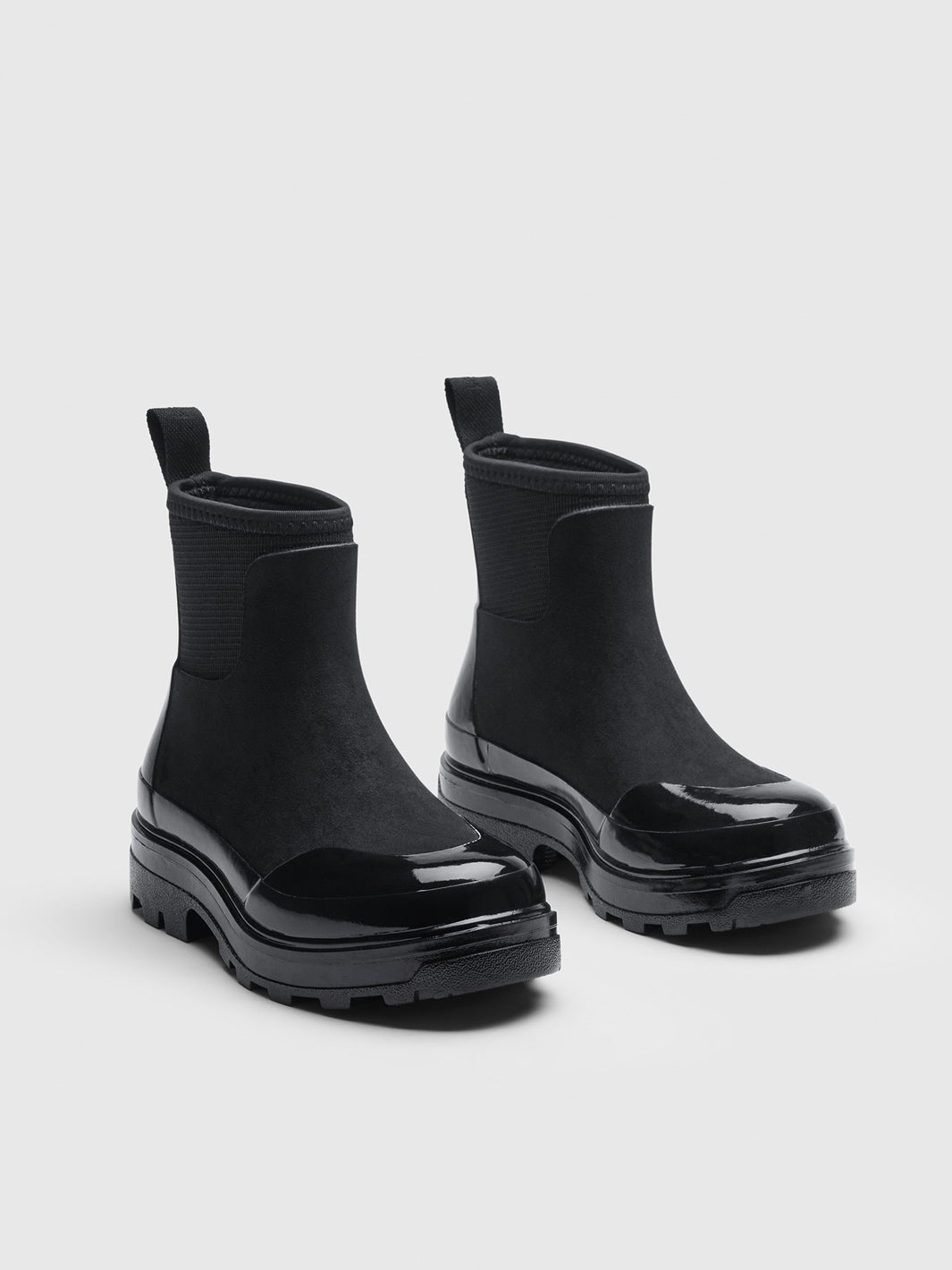 A pair of black Chelsea-style ankle rain boots with a glossy patent leather toe cap and thick lug soles.