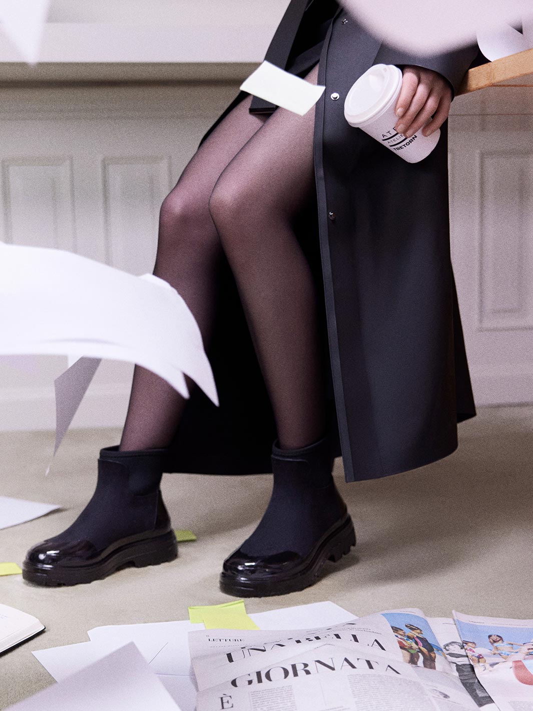 A model seated indoors wearing a pair of chunky black Chelsea-style rubber ankle rain boots and a long dark coat.
