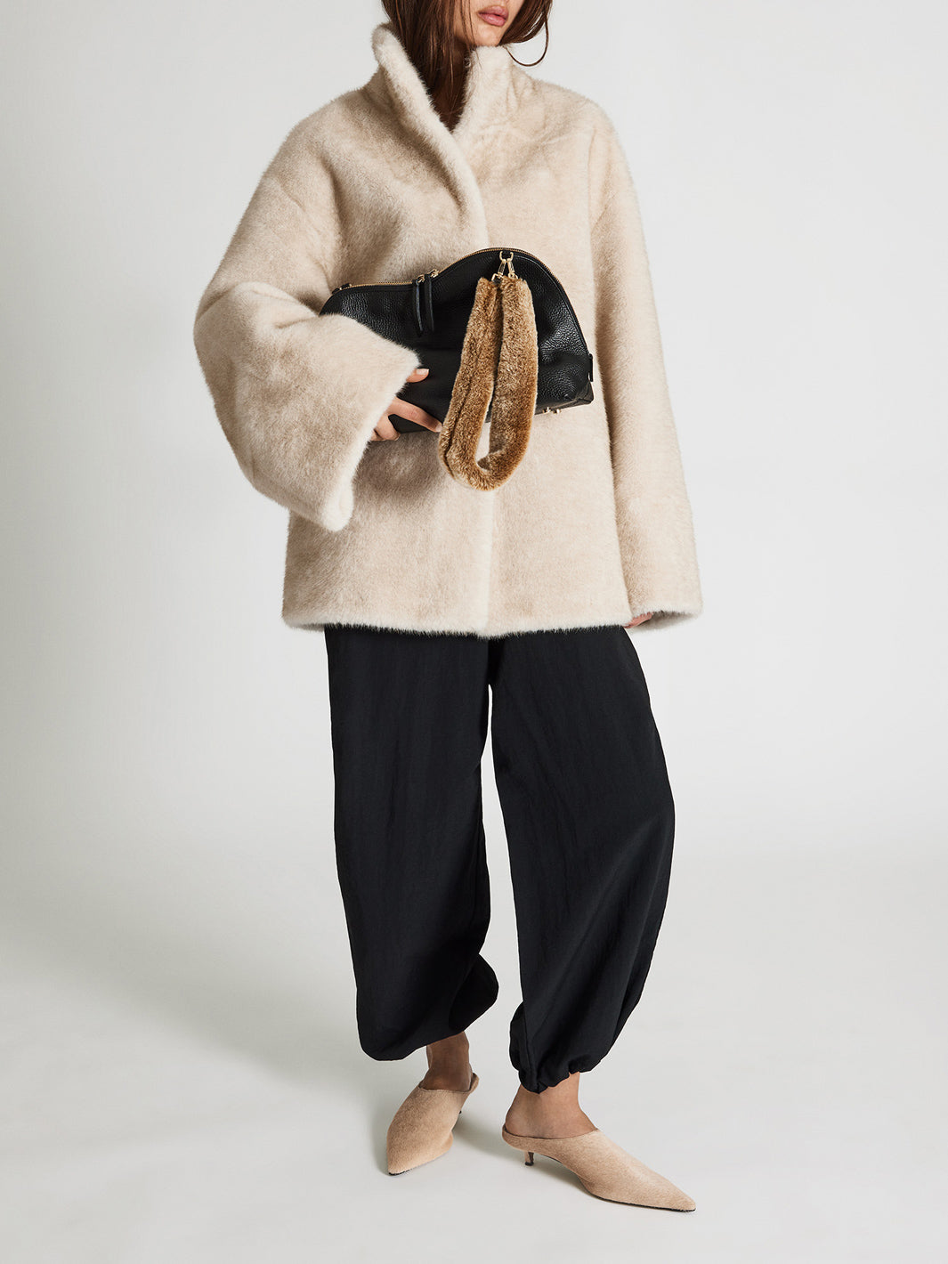 A model wearing a beige fuzzy faux-fur coat with wide sleeves, black wide-leg pants, and beige pointed kitten heels pumps, holding a black handbag with a brown fur strap against a light gray studio background.