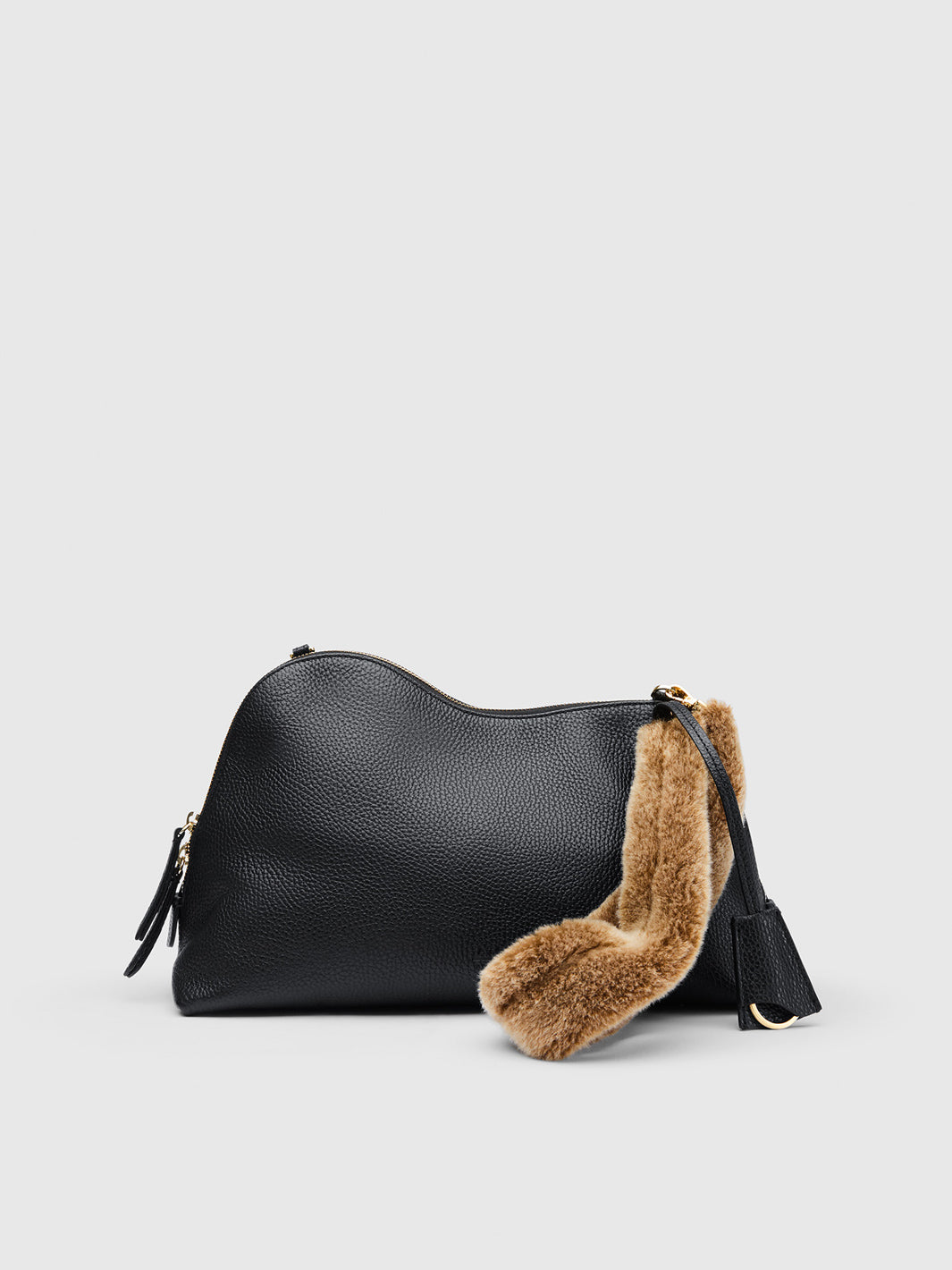 A black grained leather shoulder bag with a curved silhouette, featuring a brown fur trim along the zipper and a matching brown fur tassel charm, set against a light gray background.