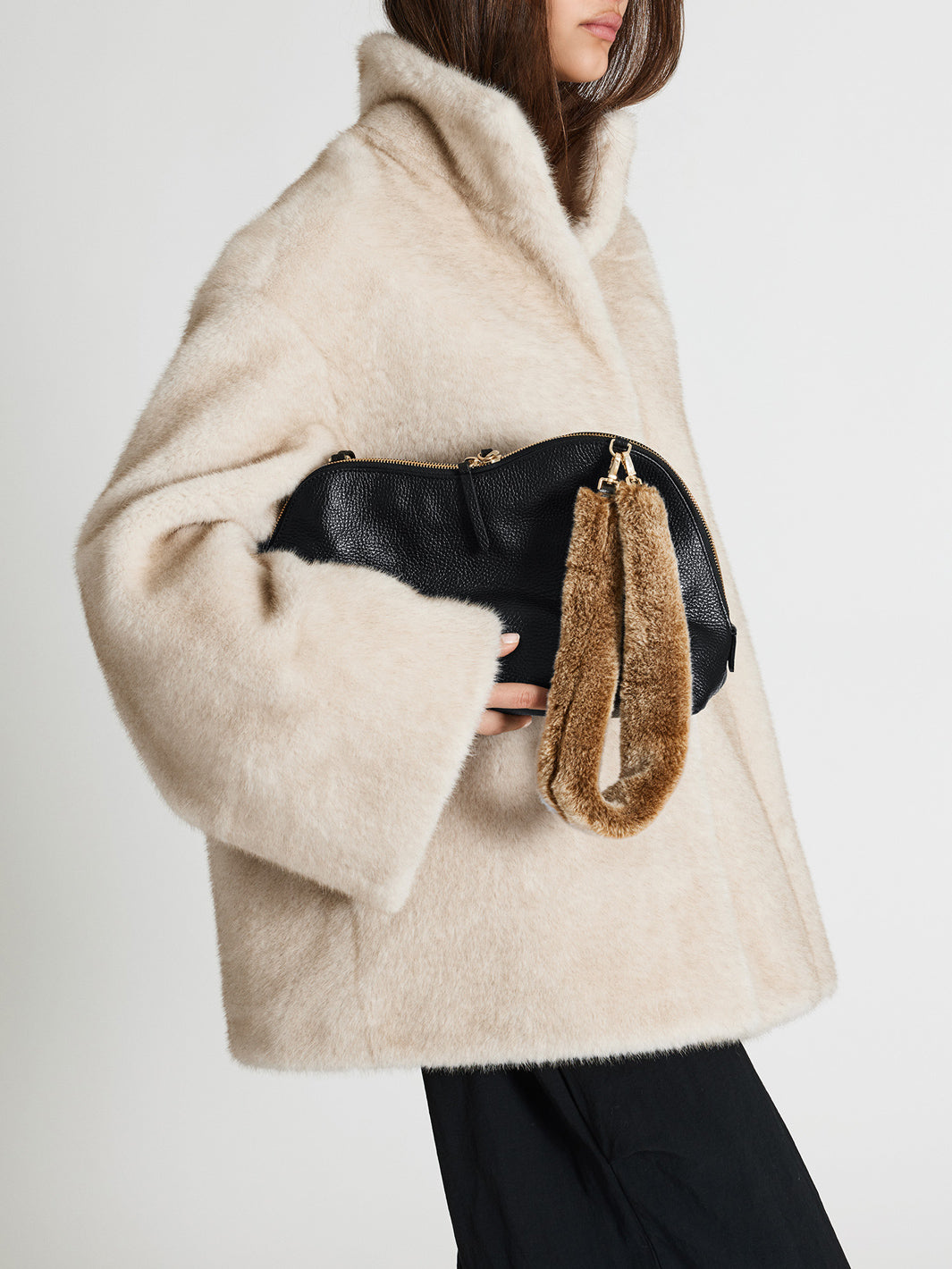 A model wearing a beige fur coat holds a black leather clutch bag with a brown fur wristlet strap and gold hardware, set against a plain light gray background.