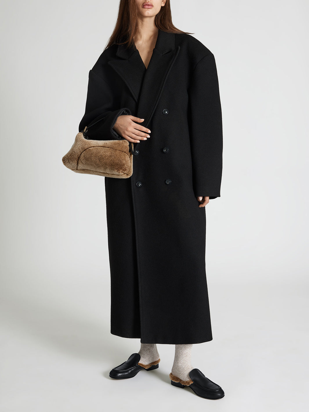 A model wearing a long black double-breasted coat, holding a brown textured shoulder bag, paired with white socks and black slip-in loafers, against a light gray studio background.